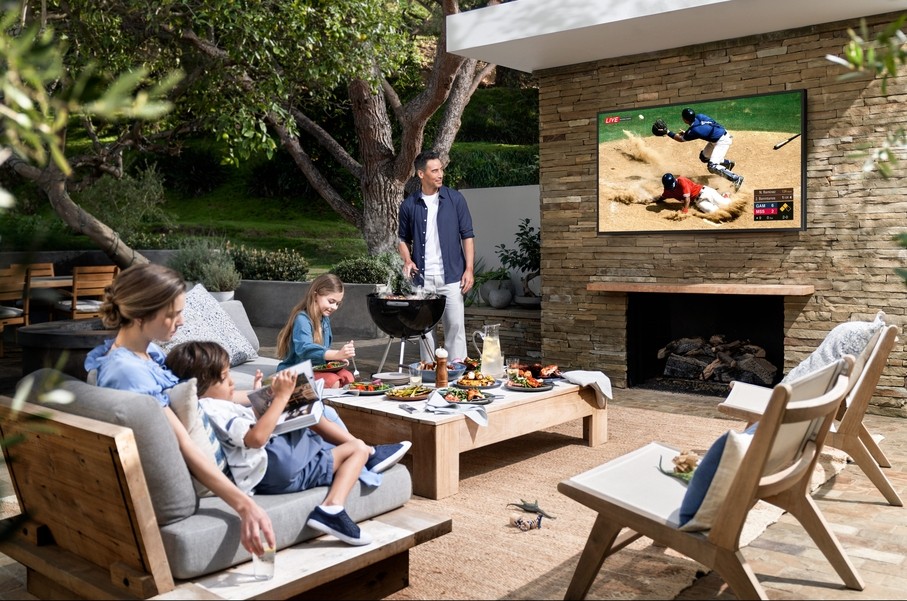 A family sitting on the patio watching a baseball game on the TV while dad makes food on a small grill. 